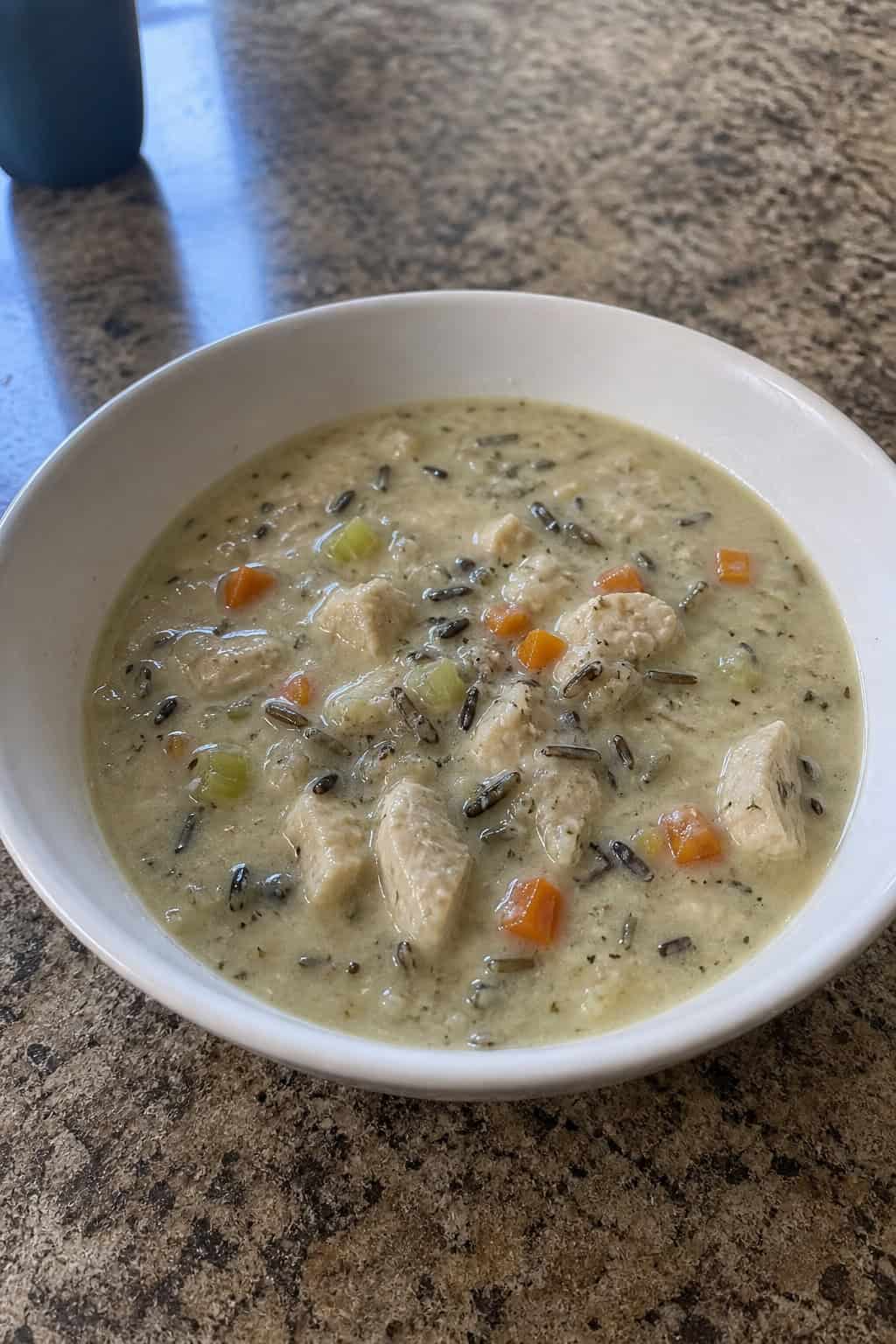 A bowl of creamy chicken and wild rice soup garnished with fresh thyme on a wooden table with a spoon and napkin nearby.