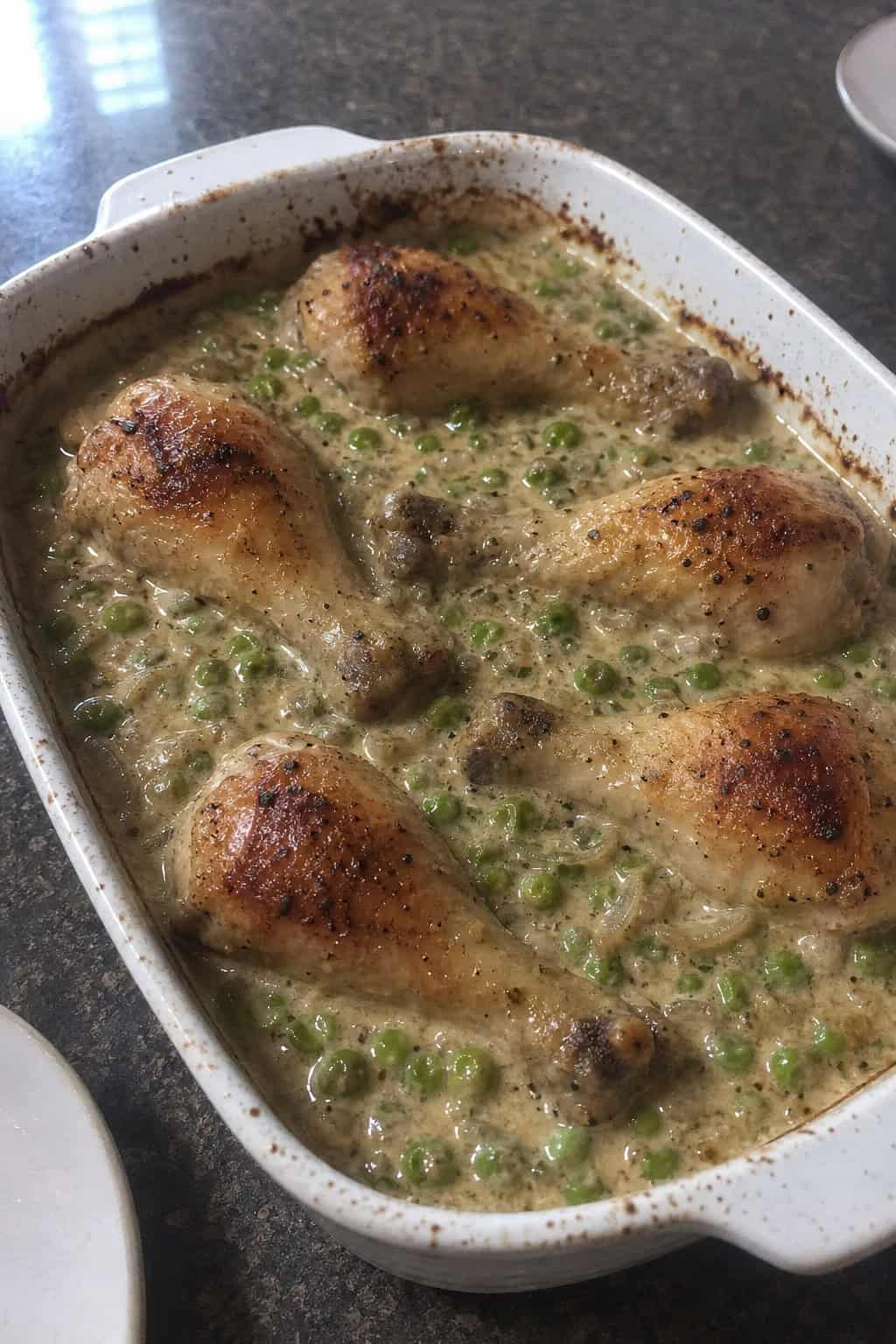 A golden-topped chicken leg casserole with visible peas and a creamy sauce served in a deep rustic ceramic baking dish on a wooden table.