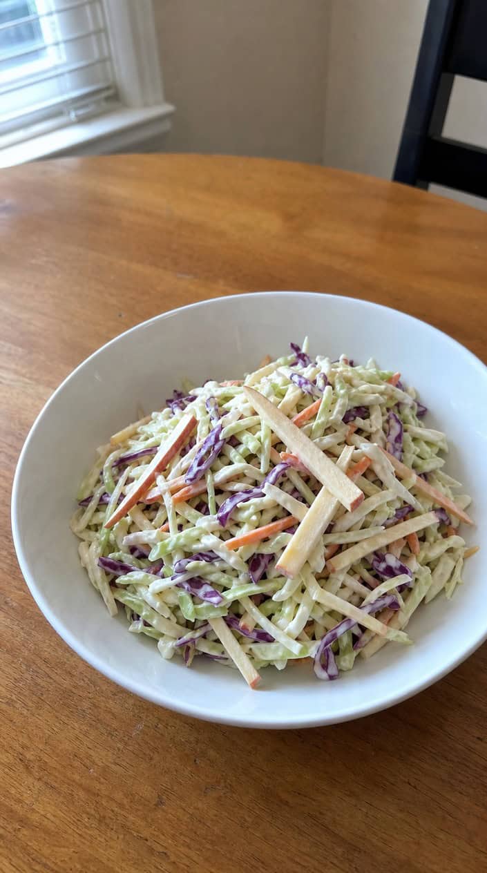 A bowl of creamy coleslaw with shredded cabbage and apple slices on a wooden table outdoors, with summer greenery in the background.