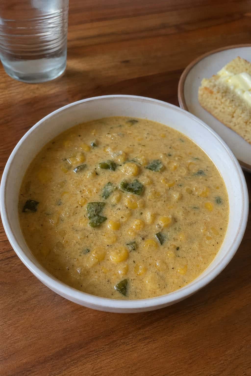 A bowl of creamy corn and poblano chowder garnished with herbs on a wooden table with fresh corn and poblano peppers nearby.