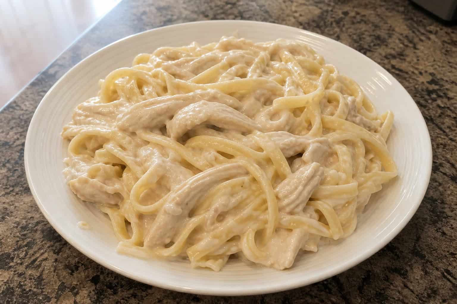 Bowl of creamy chicken Alfredo pasta garnished with parsley on a wooden table with a slow cooker in the background.
