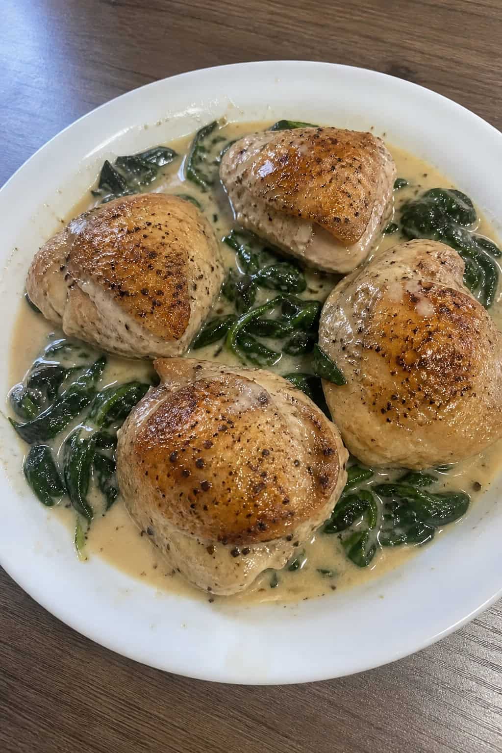 Creamy Dijon chicken thighs on a wooden table with a pale golden cream sauce pooling around them, fresh herb sprigs alongside and a small sauce bowl just visible at the edge.