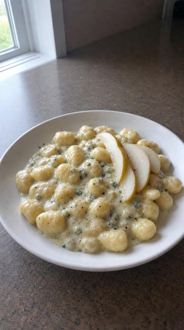 Pale ivory gnocchi coated in a creamy Gorgonzola sauce with translucent pear slices and scattered flat-leaf parsley on a wide shallow bowl over a linen-textured wooden surface.