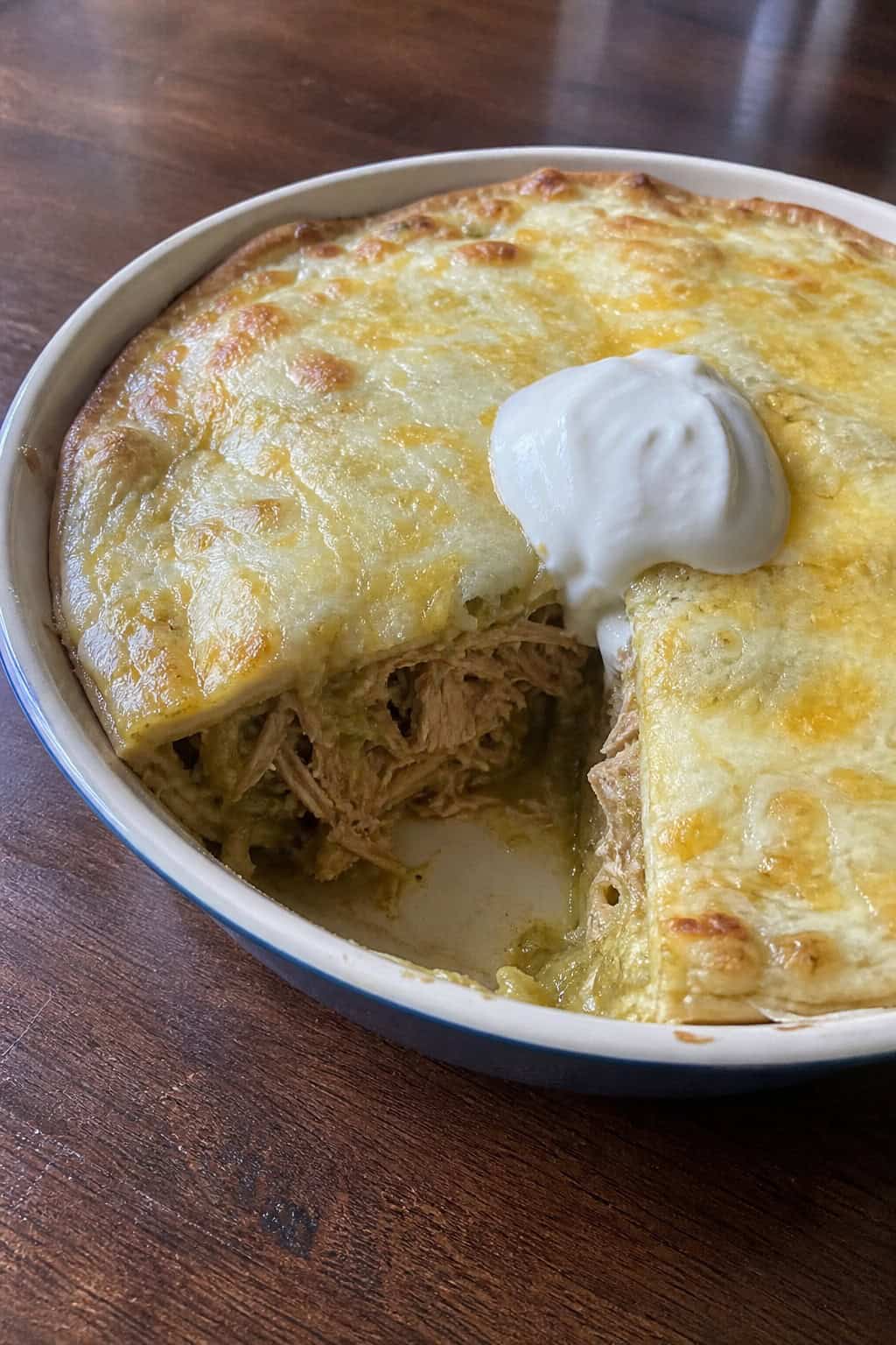 A sliced creamy green chile pork enchilada pie in a round baking dish, topped with a white sour cream drizzle and fresh cilantro, with roasted green chiles and lime wedges on the wooden table.