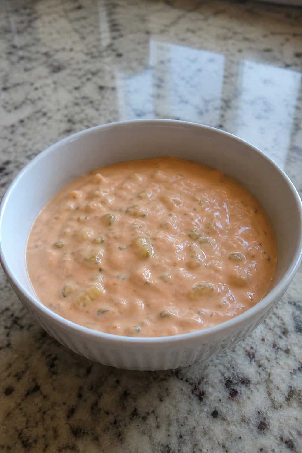 A bowl of creamy Thousand Island dressing garnished with sweet pickle relish and paprika, placed on a wooden surface with fresh pickles and spices around it.