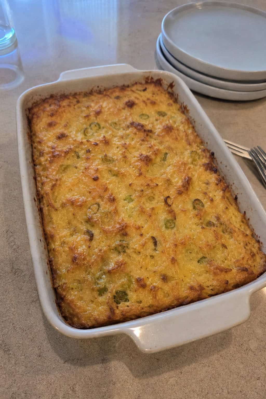 A baked hashbrown casserole with melted cheese and leeks in a ceramic dish on a wooden table with fresh ingredients around it.