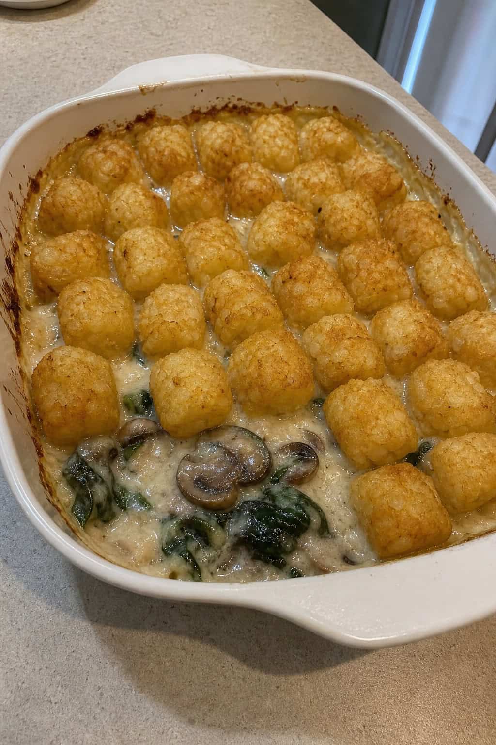 A freshly baked creamy mushroom and spinach tater tot casserole in a round ceramic dish on a wooden table, with the golden tots slightly overlapping and a dark green spinach filling visible at the rim.