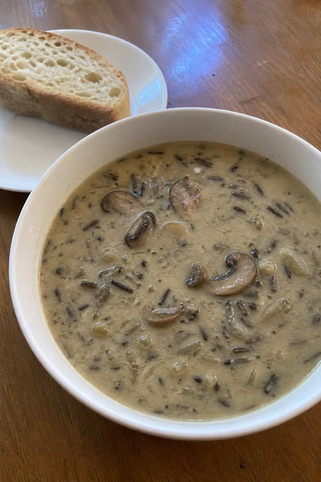 A steaming bowl of creamy mushroom and wild rice soup on a wooden table with fresh herbs and bread nearby.