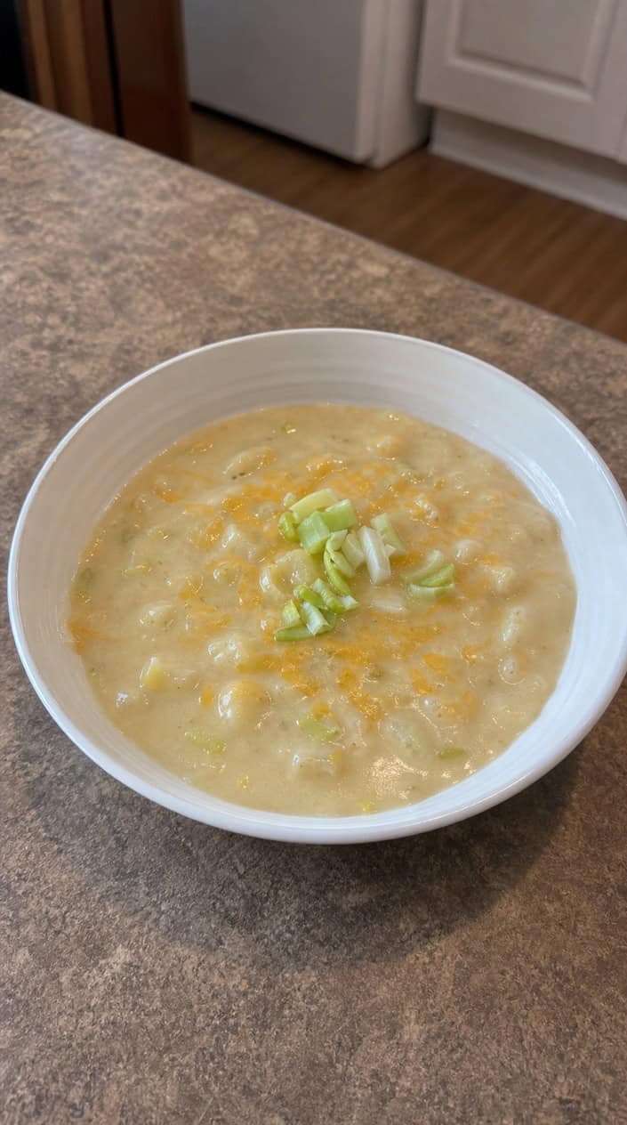 A bowl of creamy potato soup garnished with cheddar cheese and chopped leeks on a wooden table with fresh potatoes and leeks nearby.