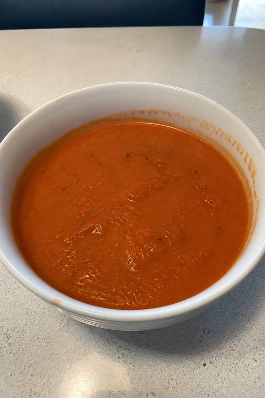 A bowl of creamy roasted tomato and basil soup garnished with fresh basil on a wooden table surrounded by ingredients like tomatoes and garlic.