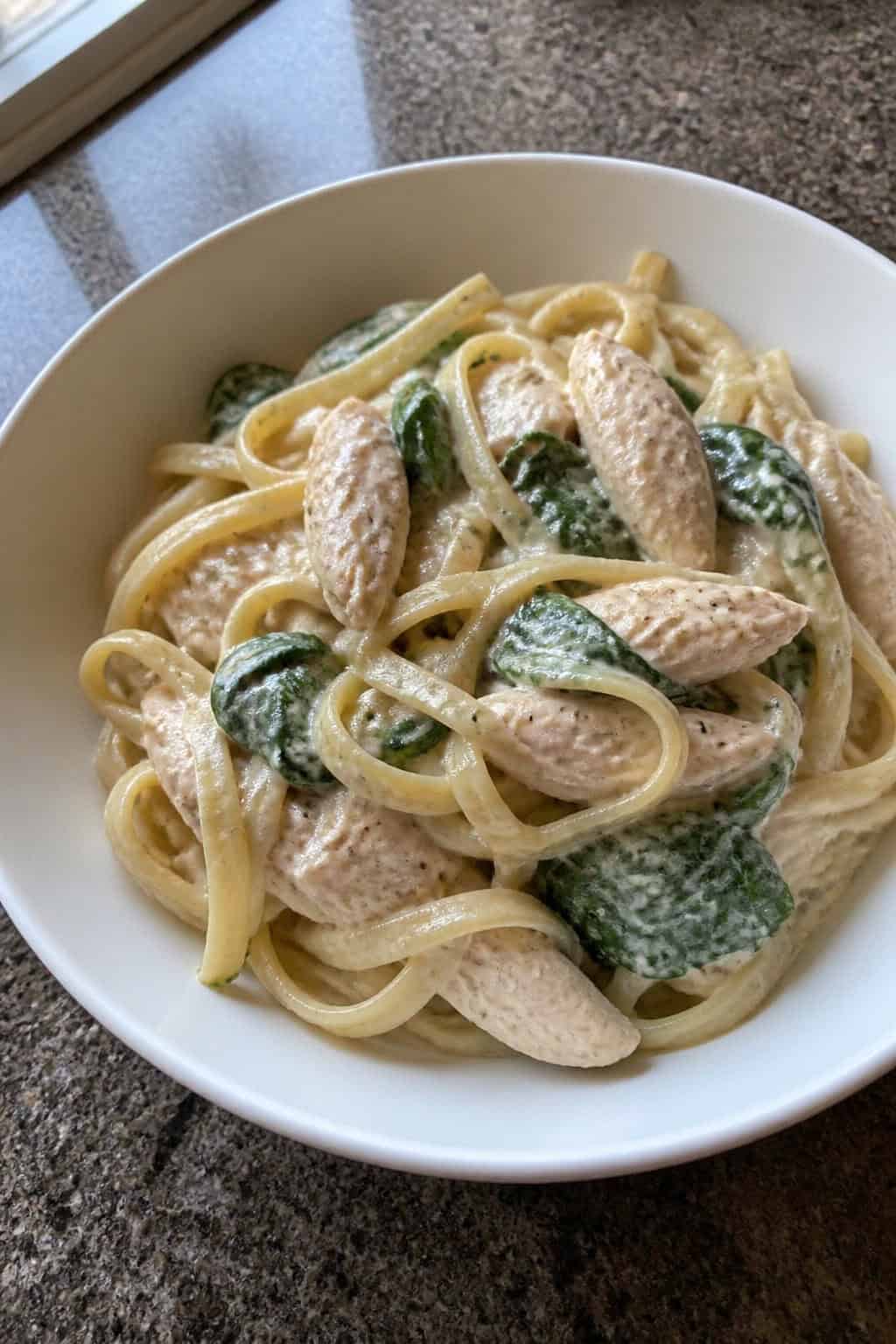 A wide pasta bowl on a wooden table filled with penne in a creamy Parmesan sauce, shredded chicken, and wilted dark green spinach leaves.