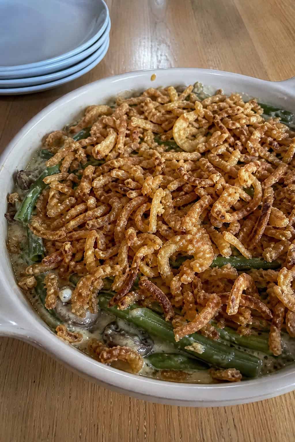 A creamy green bean casserole topped with a generous layer of crispy fried onions in a rustic oval baking dish, with fresh green beans and sliced mushrooms arranged nearby on a wooden surface.