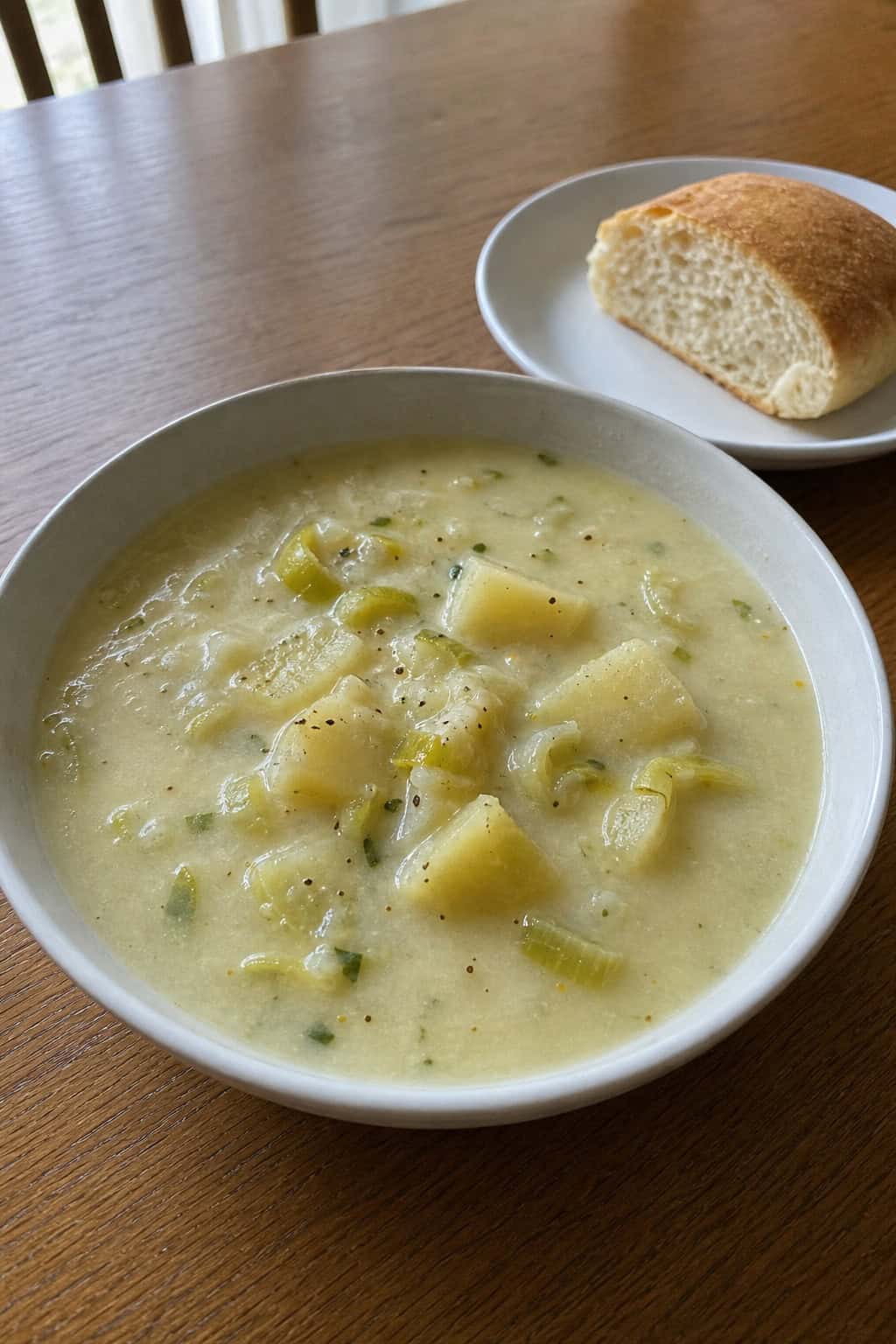 A bowl of creamy potato leek soup garnished with chopped chives on a wooden table surrounded by fresh leeks and potatoes.