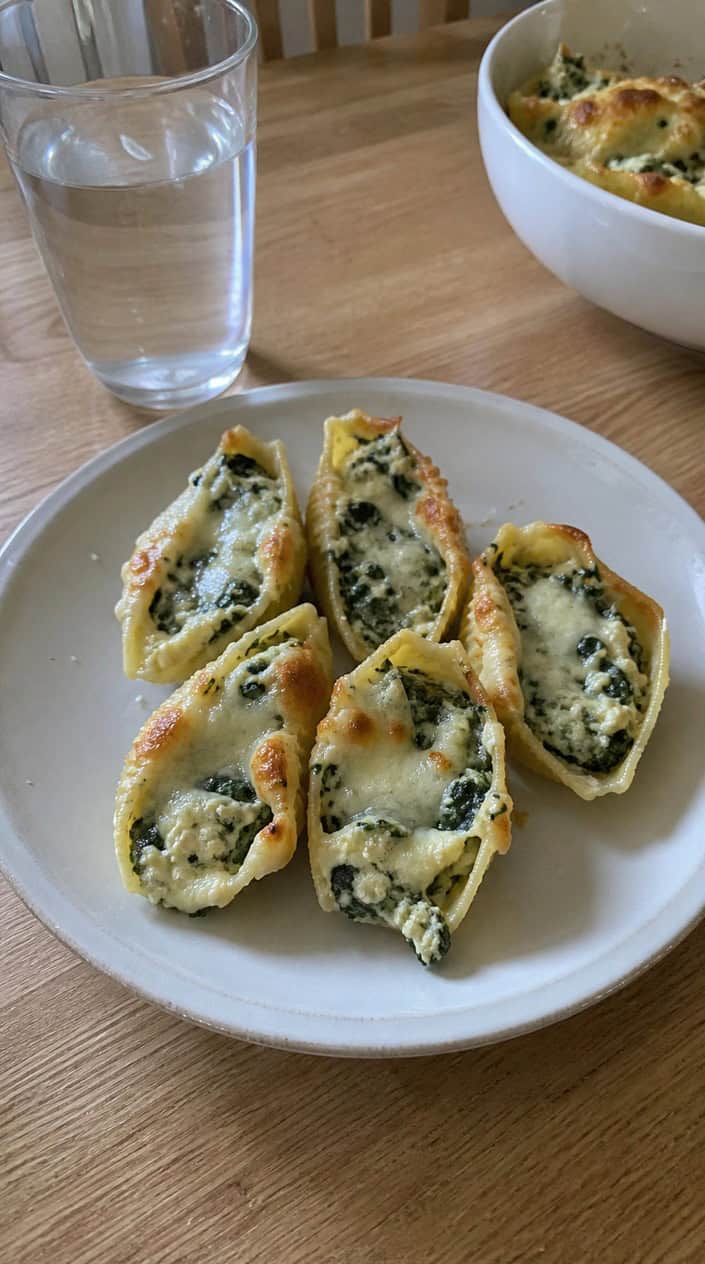 Plate of creamy spinach and ricotta stuffed pasta shells on a wooden table with a glass of water and kitchen items in the background.