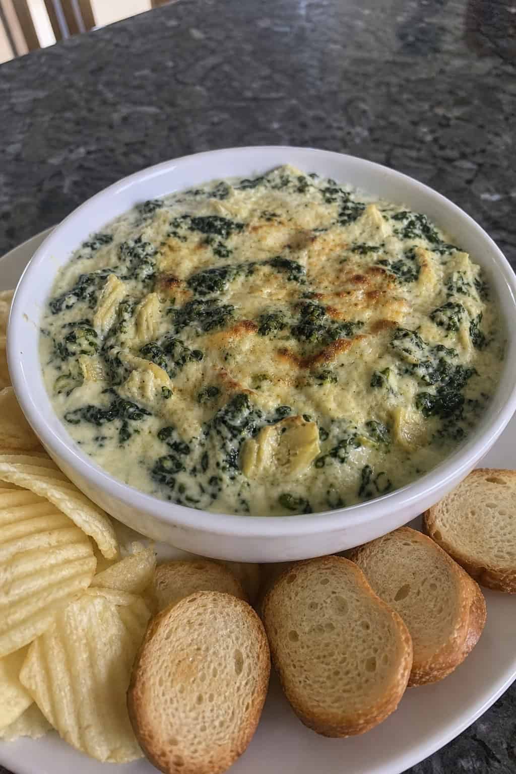 A bowl of creamy spinach-artichoke dip with vegetable sticks and toasted bread slices on a wooden board.