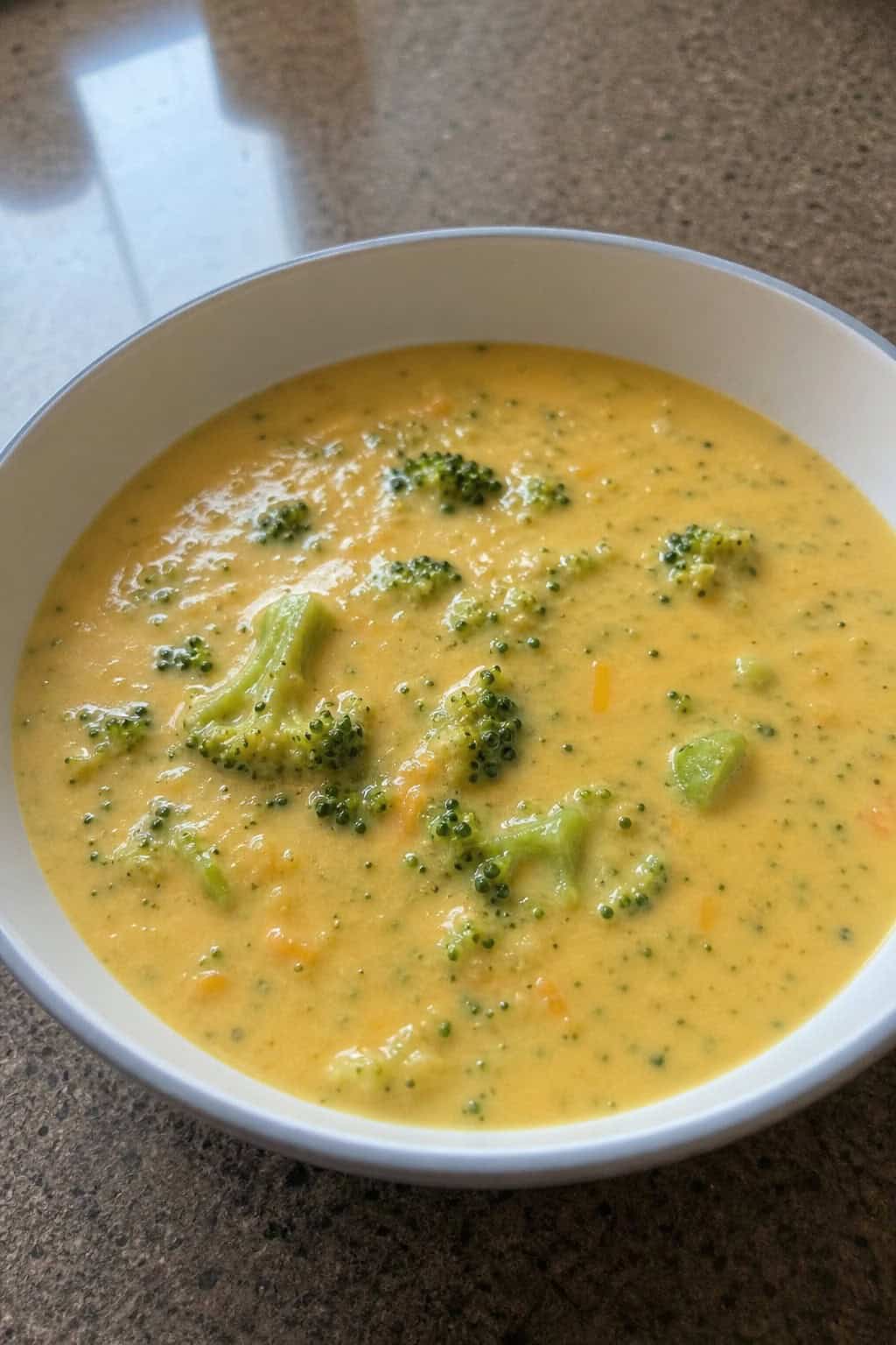 A bowl of creamy broccoli cheddar soup with broccoli florets and melted cheese on a wooden table, surrounded by fresh broccoli and a wedge of cheddar cheese.
