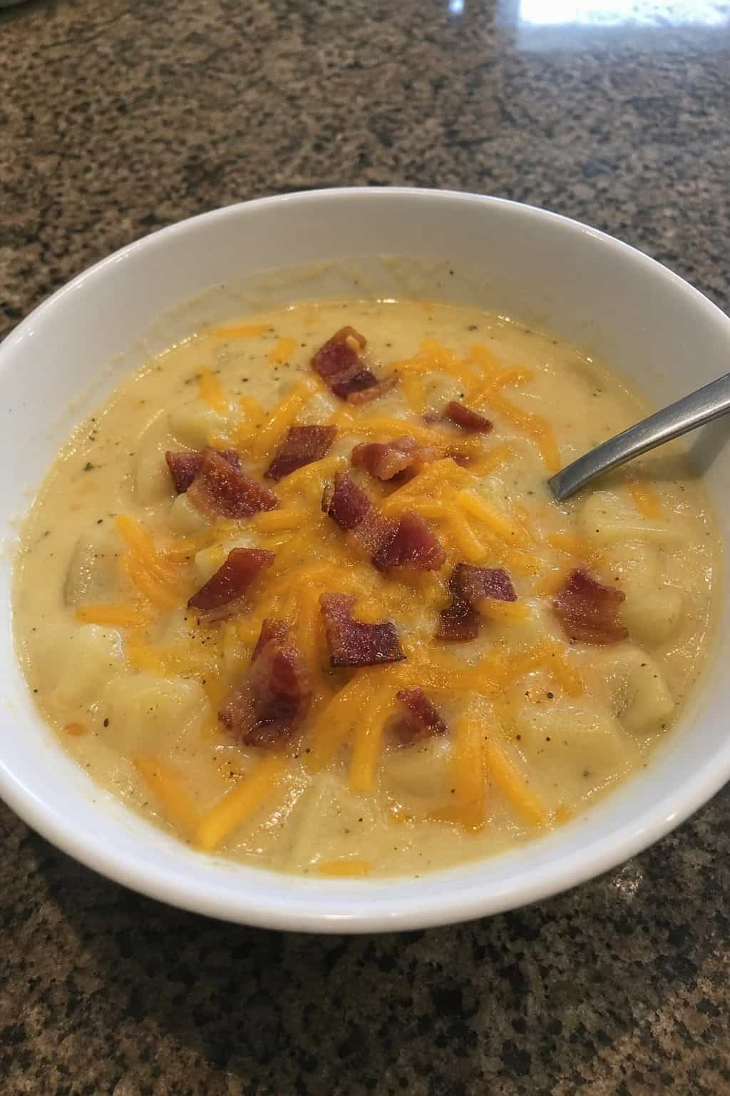 A bowl of creamy potato soup topped with bacon and cheddar cheese on a wooden table with fresh ingredients around it.