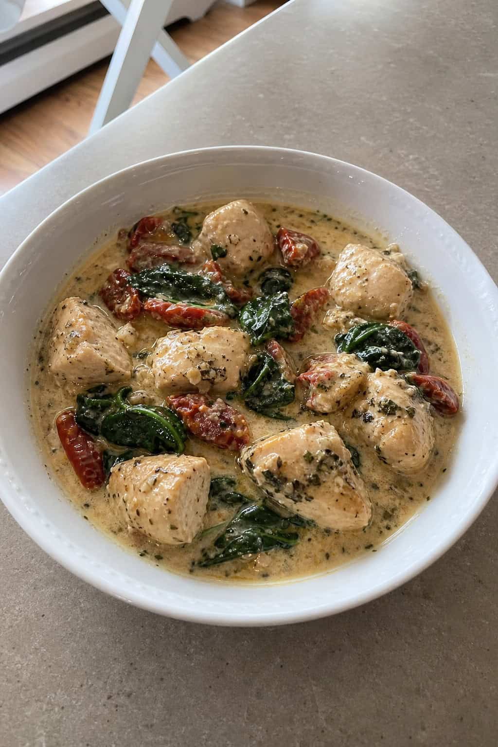 A bowl of creamy Tuscan chicken with sun-dried tomatoes and spinach on a wooden table with fresh ingredients around it.