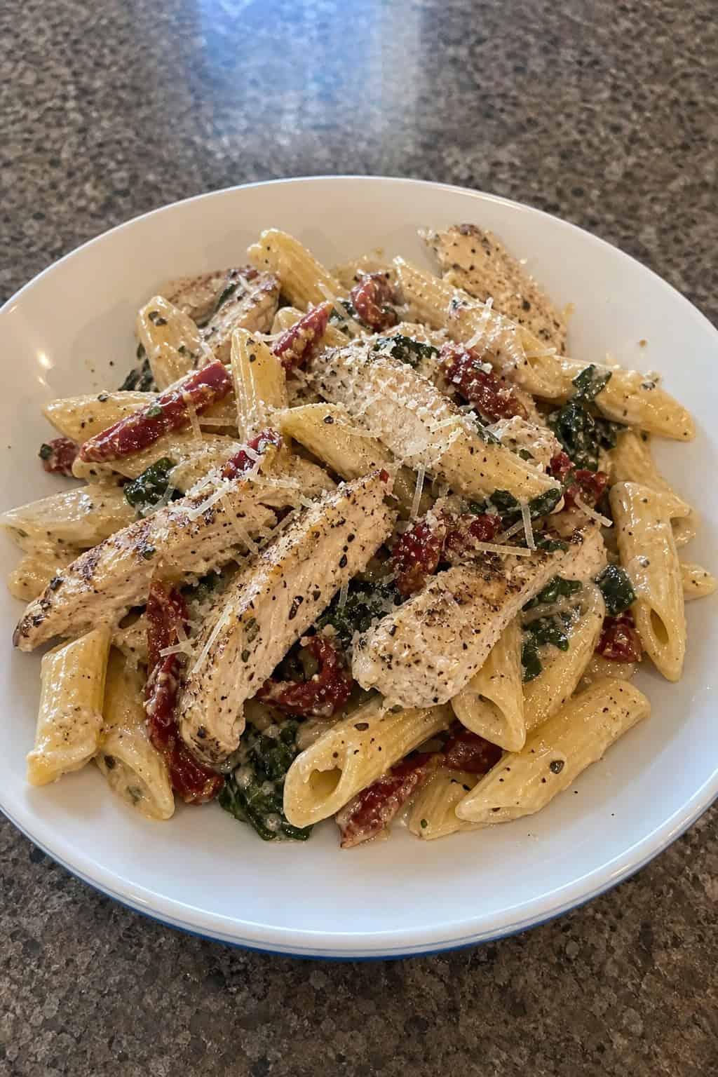 A bowl of creamy Tuscan chicken penne pasta with spinach, sun-dried tomatoes, and grilled chicken on a wooden table.