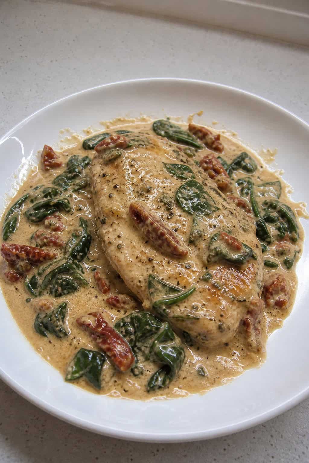 A plated creamy Tuscan chicken breast with sun-dried tomatoes and spinach, garnished with basil and Parmesan cheese.