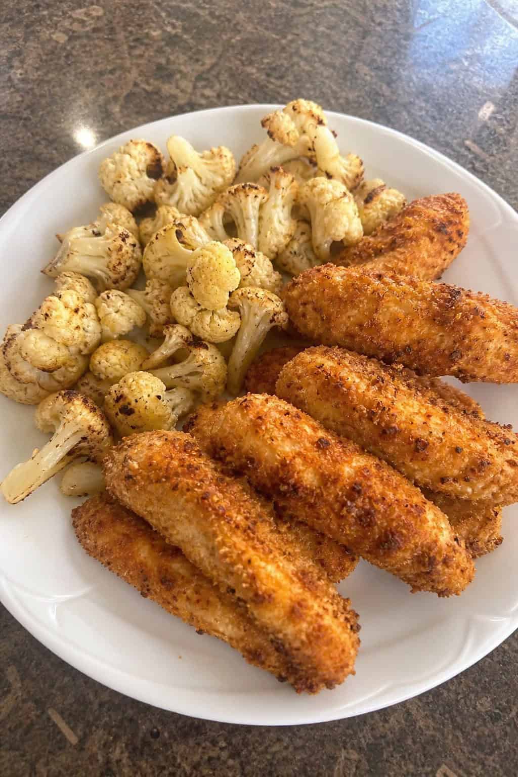 A plate of crispy baked chicken tenders served with roasted cauliflower and garnished with fresh herbs.