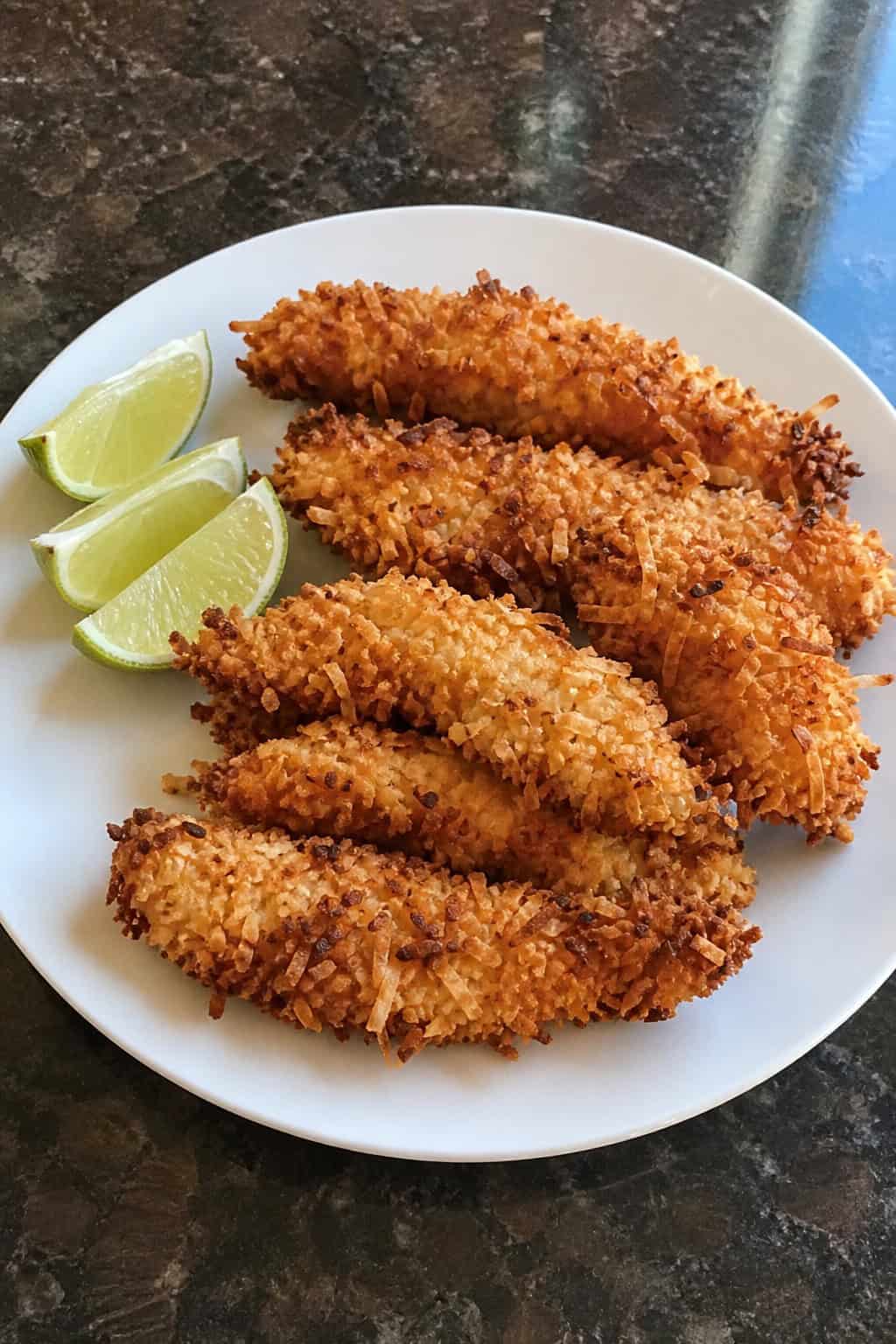 Crispy coconut-crusted chicken tenderloins arranged on a white plate with fresh lime wedges on a wooden surface.