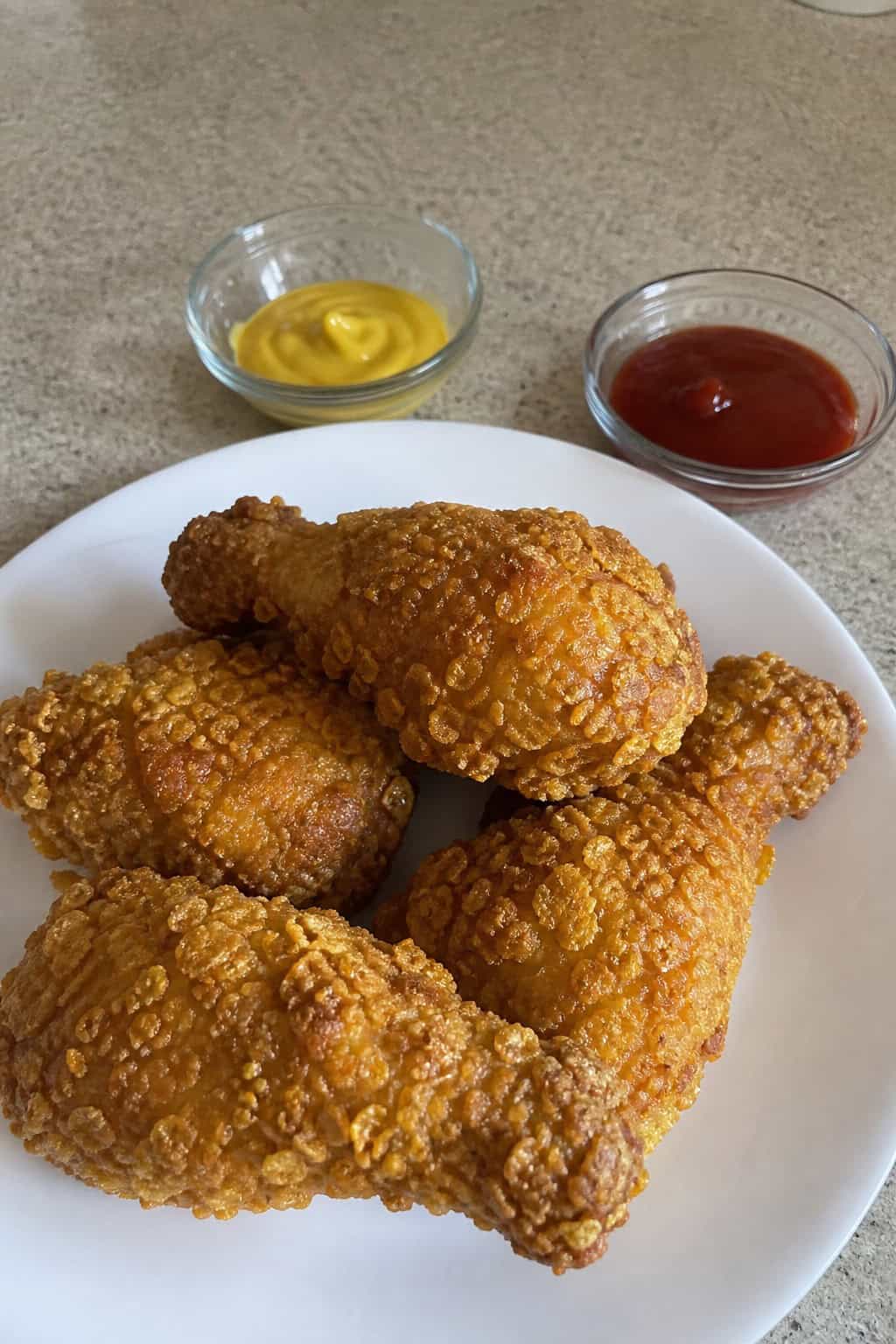 Crunchy cornflake-coated chicken drumsticks on a wooden board with visible flaky golden crust, fresh herb garnish, and small dipping sauce bowls.