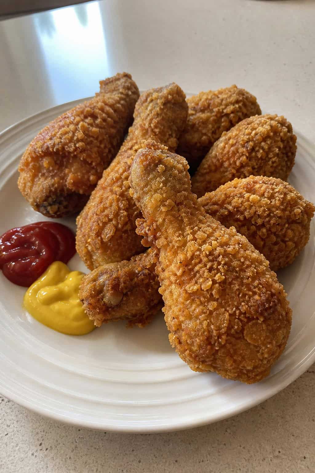 Shatteringly crunchy cornflake-coated chicken drumsticks on a rough wooden board with a small ramekin of ketchup and a ramekin of mustard, with fresh dill scattered around.