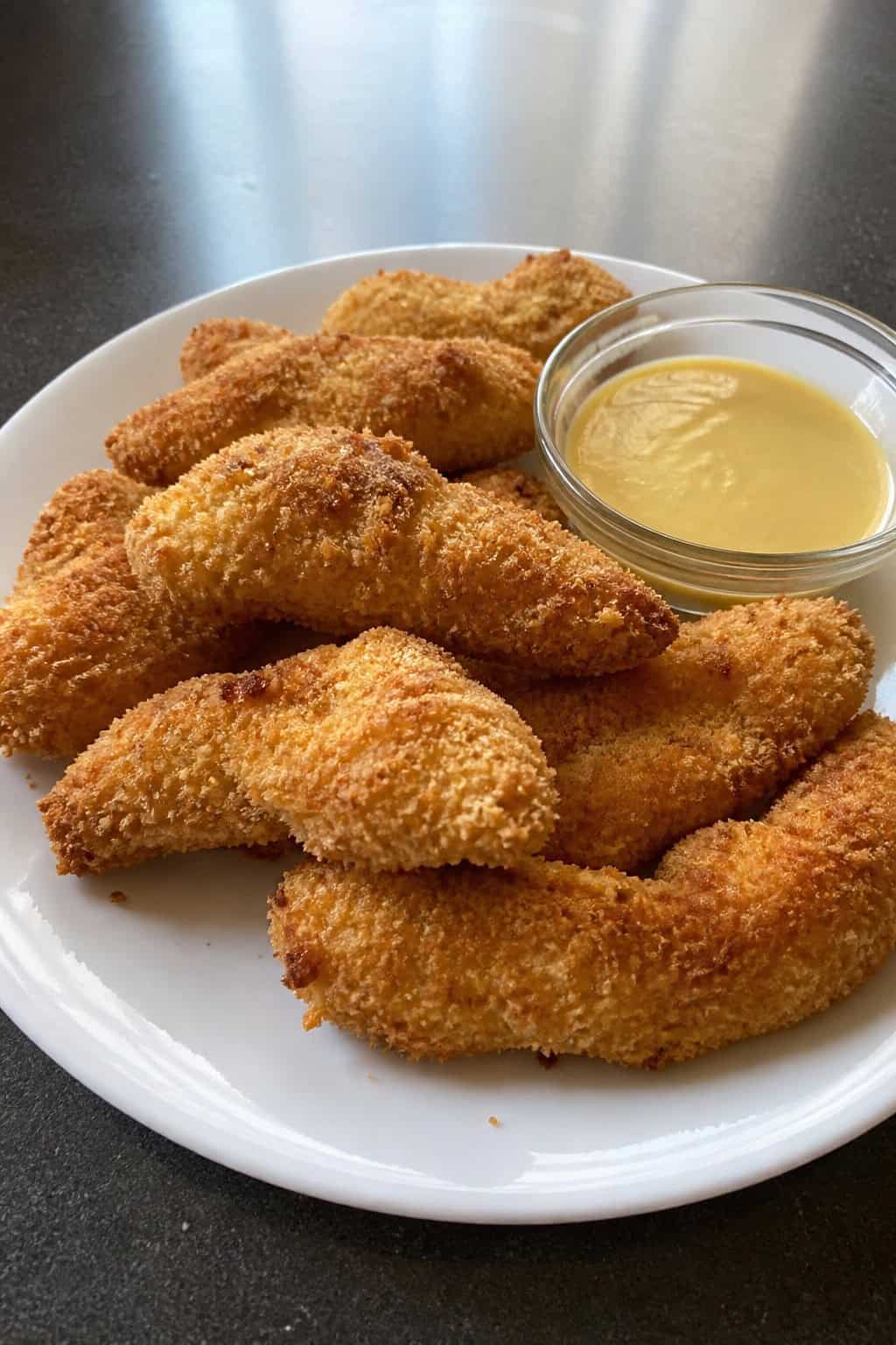 Plate of crispy oven-baked chicken tenders with a bowl of dipping sauce and fresh herbs on a wooden table.