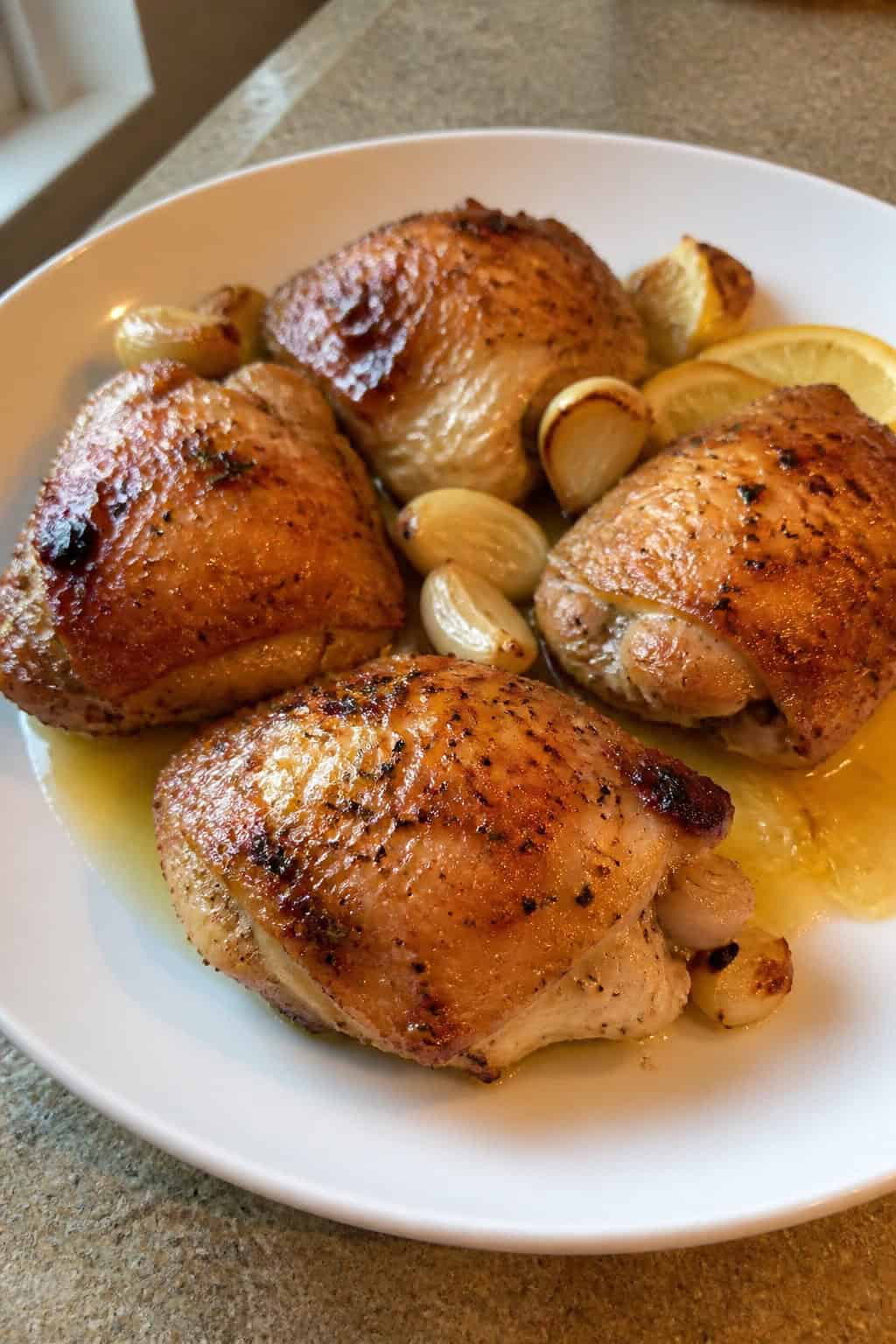 A plate of crispy oven-baked lemon garlic chicken thighs garnished with lemon slices, garlic cloves, and fresh herbs.
