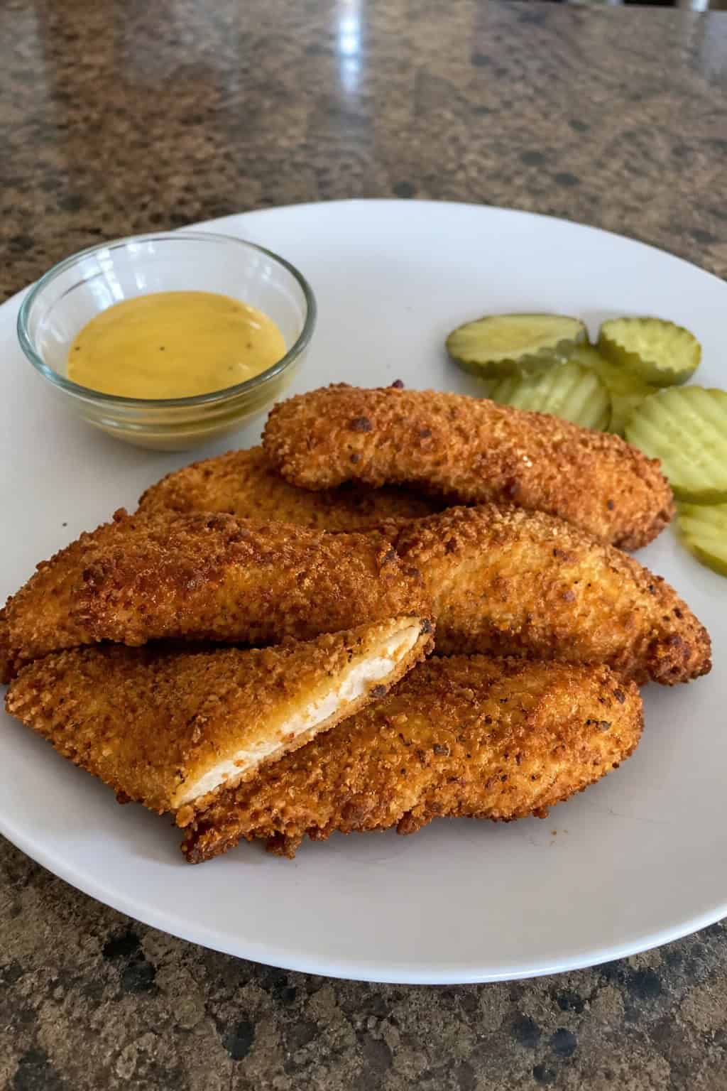 Golden crispy chicken tenders on a wooden board garnished with fresh dill, served with two small bowls of dipping sauce.