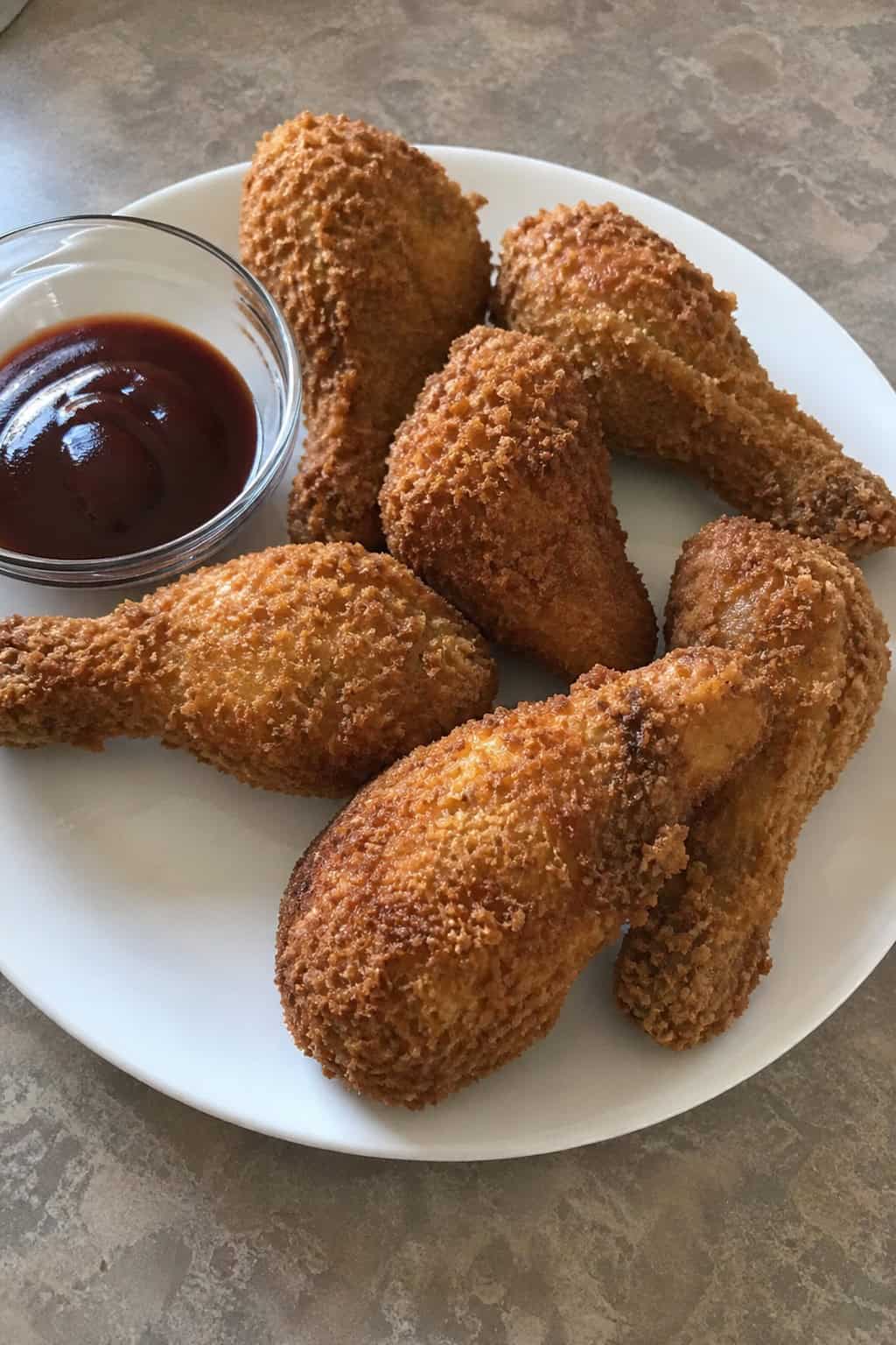 Deeply golden panko-crusted chicken drumsticks on a rough wood serving board, scattered with fresh herb leaves and two small bowls of dipping sauce.