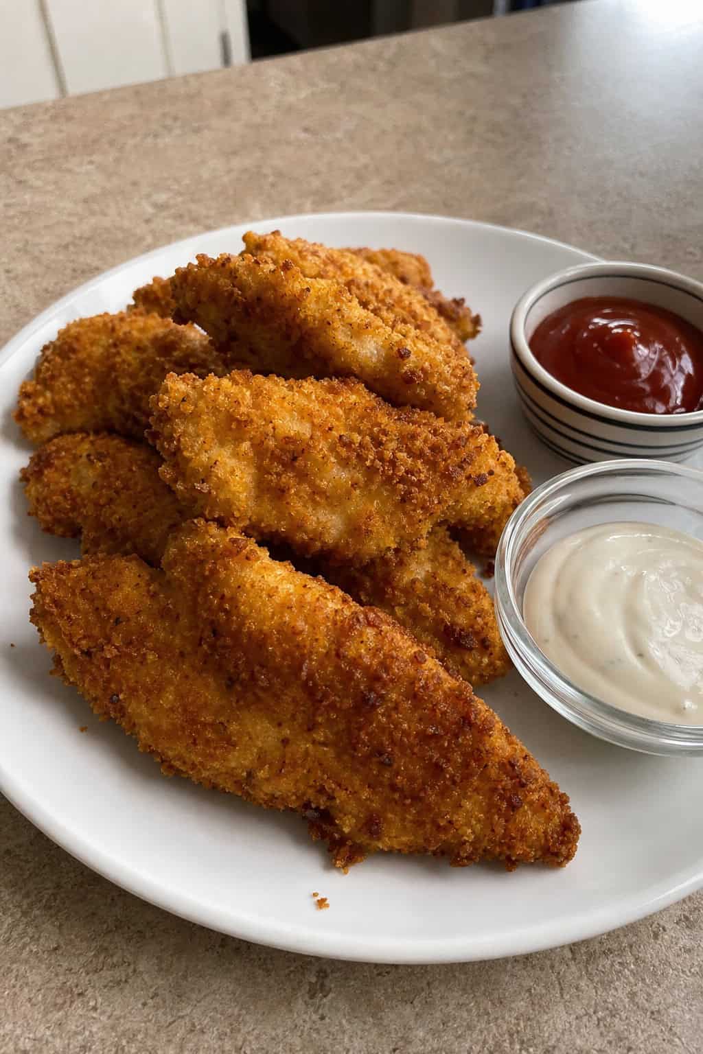 A plate of crispy golden chicken tenders garnished with parsley and served with dipping sauces on a wooden board.
