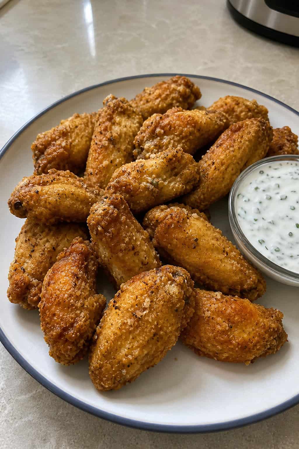 A plate of crispy ranch-coated chicken wings served with a bowl of dill and chive dip on a wooden table.