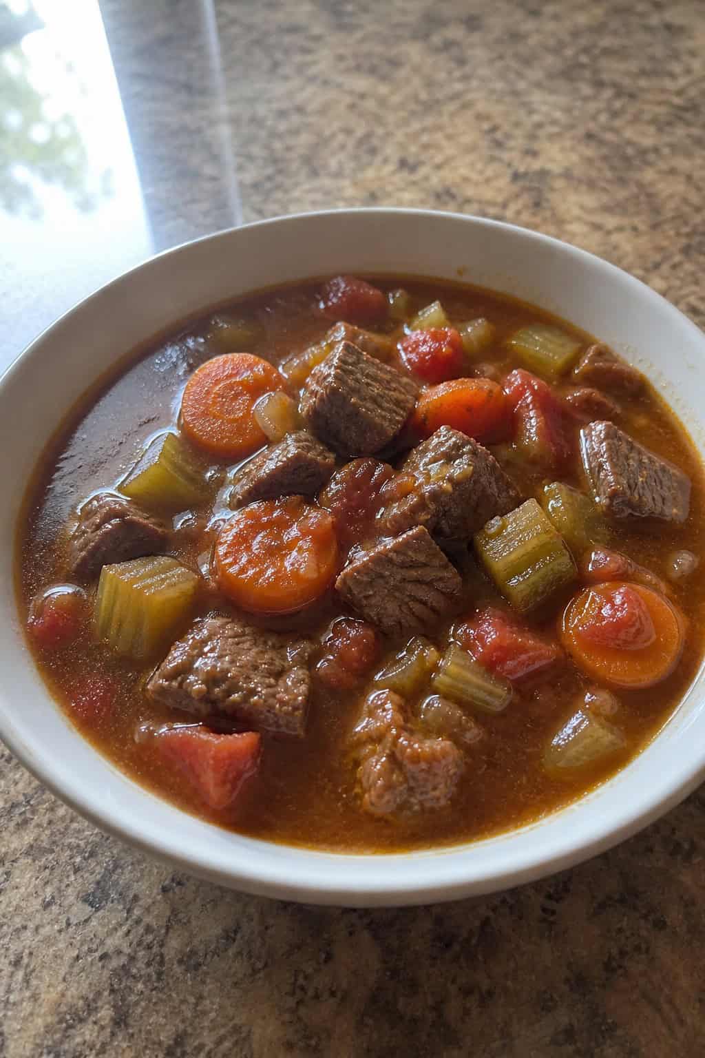 A bowl of beef and vegetable soup with fresh ingredients on a wooden table.