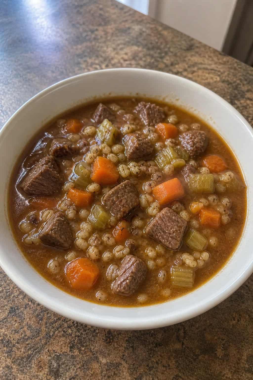 A bowl of beef barley stew with vegetables on a wooden table surrounded by fresh ingredients.