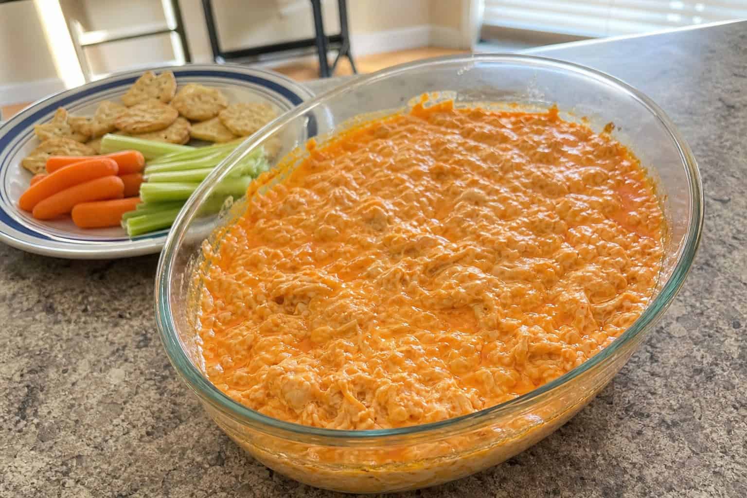 A bowl of buffalo chicken dip with vegetable sticks on a wooden board in a cozy kitchen setting.