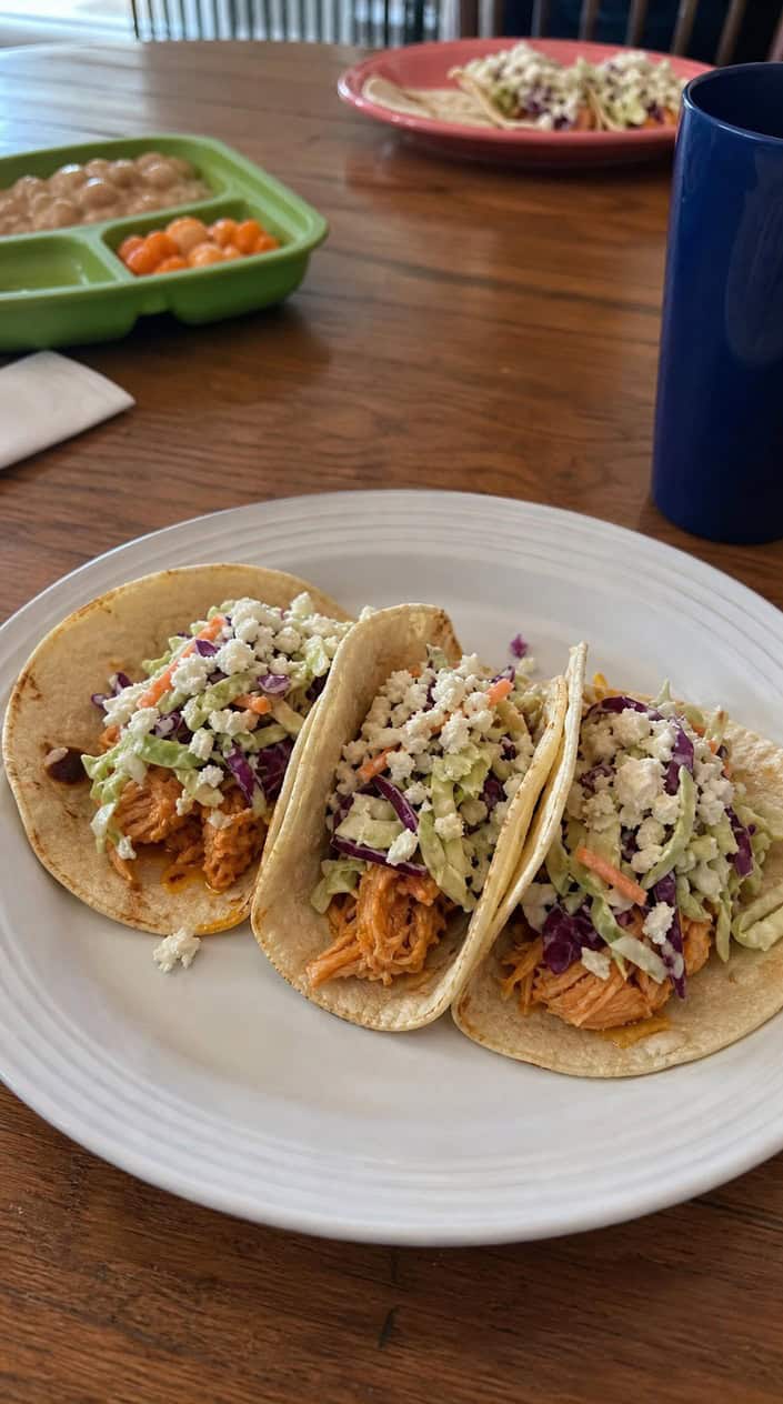 Close-up of pulled buffalo chicken tacos in small flour tortillas piled with pale shredded cabbage and blue cheese slaw on a wooden table, with lime wedges and a small bowl of dressing to the side.