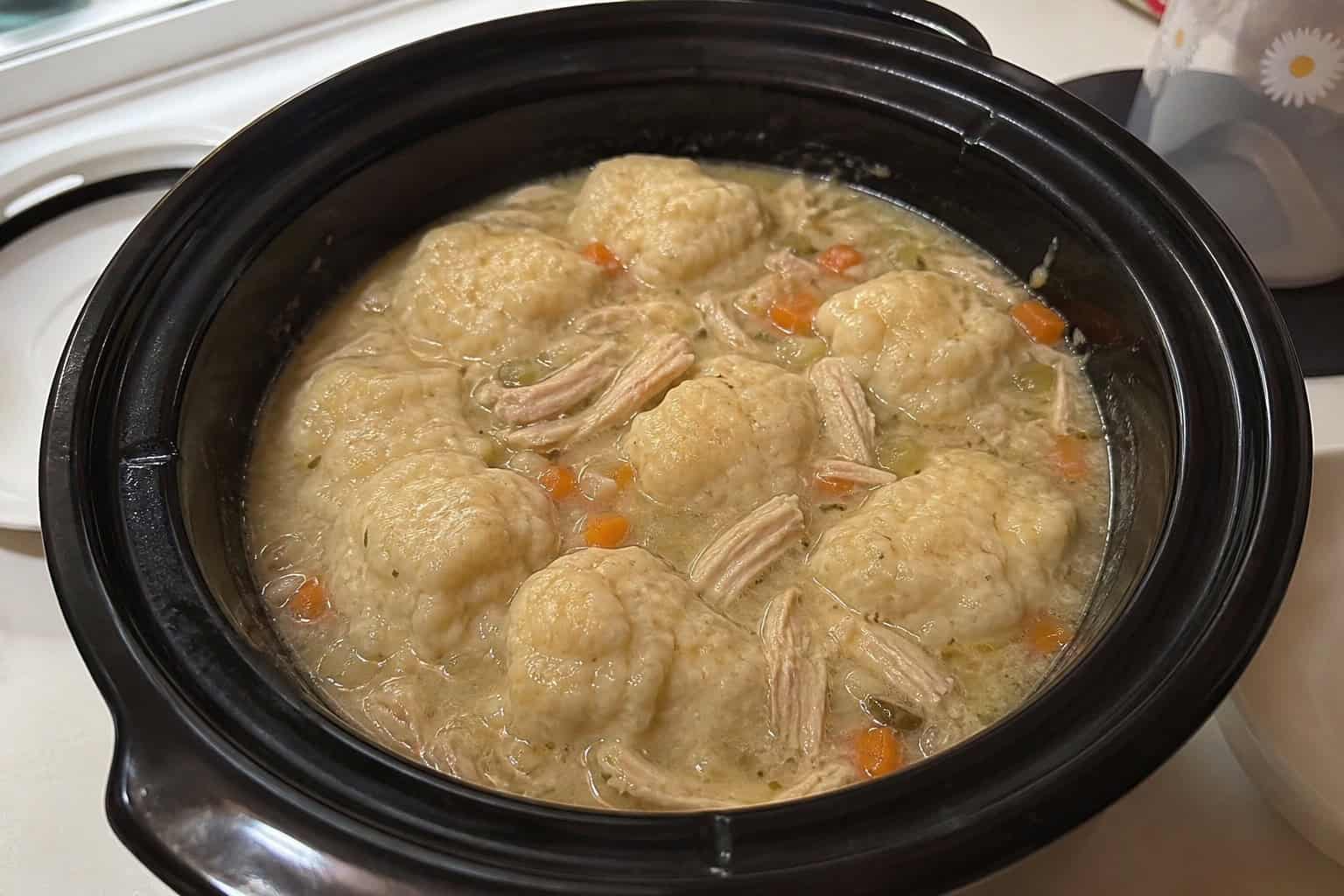A crockpot filled with steaming chicken and dumplings on a wooden countertop surrounded by fresh vegetables and herbs.