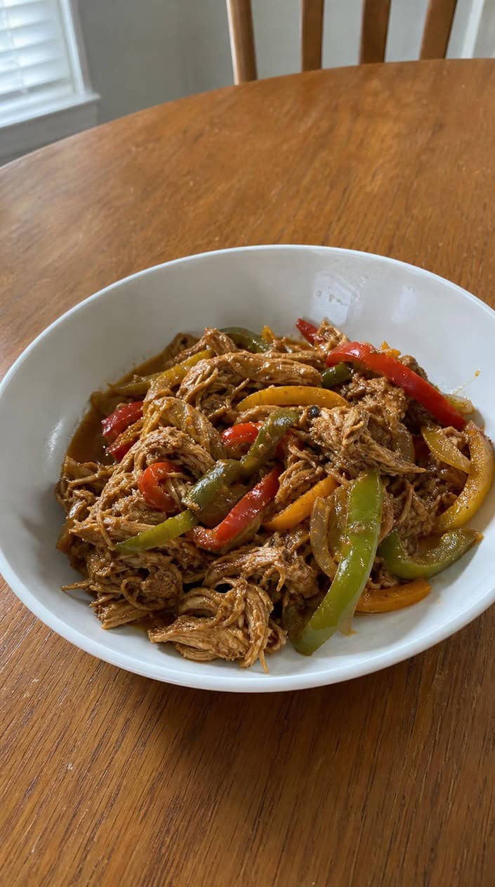 A deep bowl of saucy shredded chicken fajita filling with softened red and green bell pepper strips and caramelized onion, placed on a wooden table with fresh ingredients scattered around it.