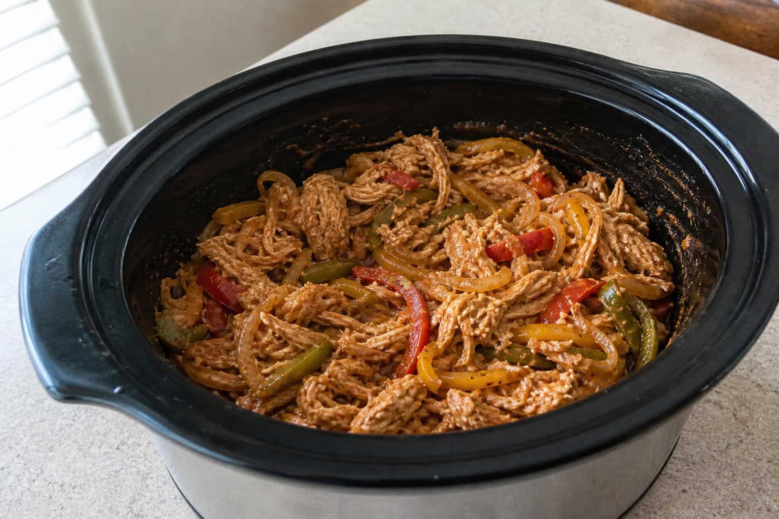 Close-up of a crockpot filled with chicken fajita filling including bell peppers and onions on a kitchen countertop with fresh ingredients nearby.