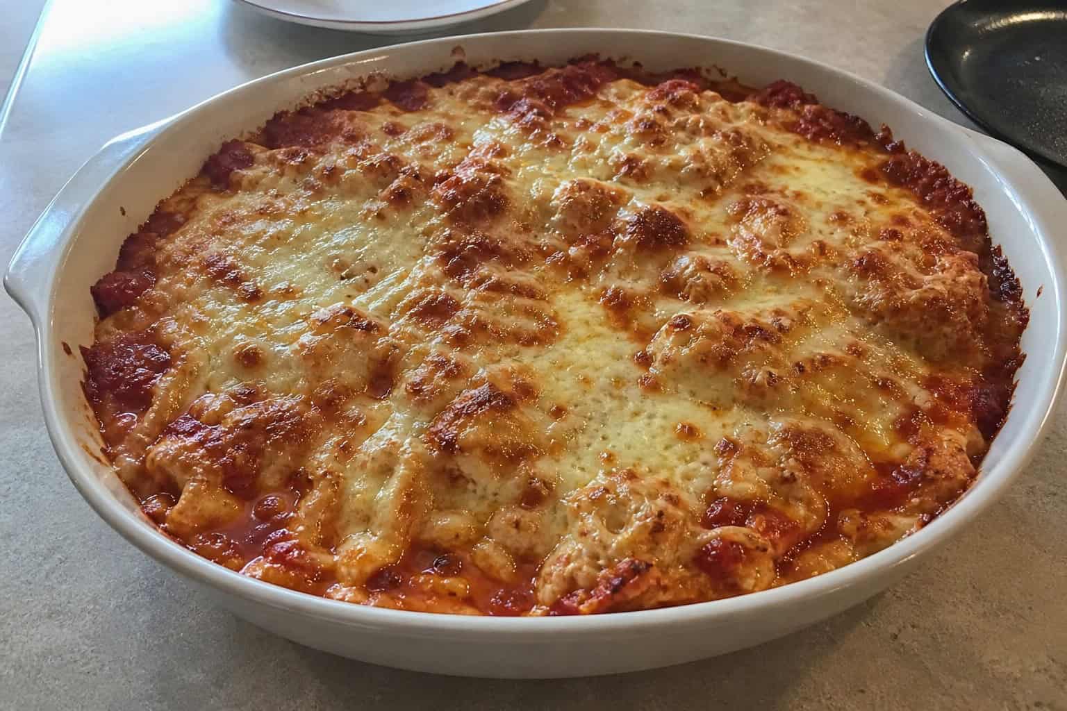 A cooked chicken parmesan casserole in a ceramic dish on a wooden table with fresh ingredients around it.