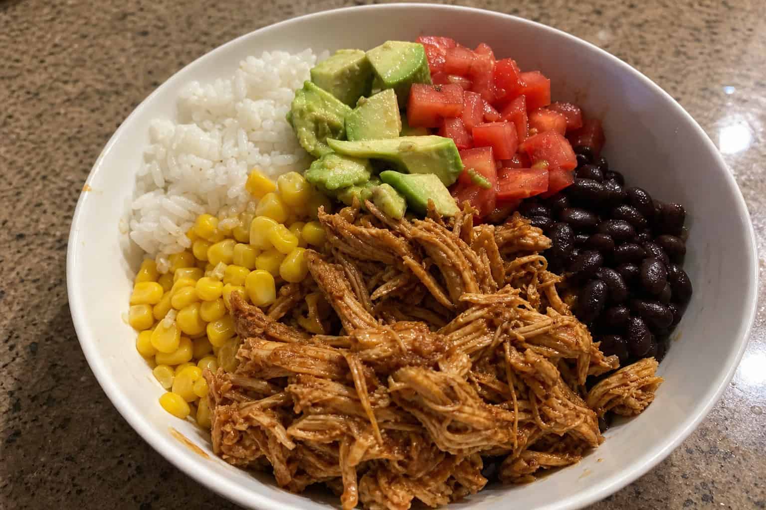 A bowl of shredded crockpot chicken taco topped with avocado, tomatoes, black beans, corn, and cilantro on a wooden table with tortilla chips and salsa nearby.