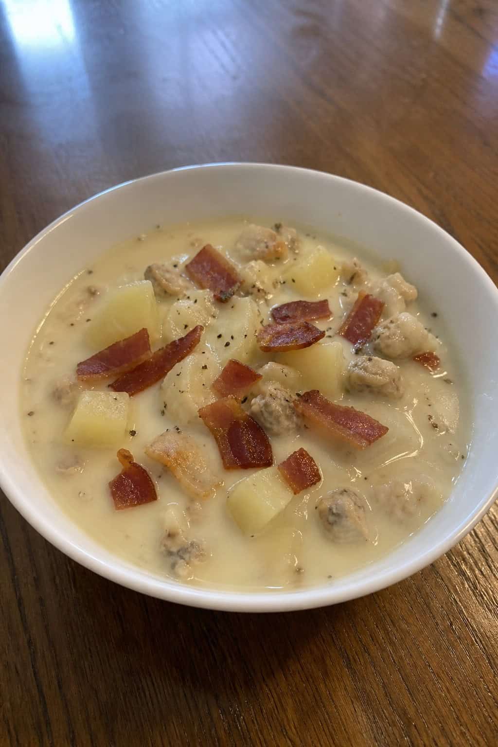 A bowl of creamy clam chowder with clams, potatoes, and crispy bacon on a wooden table with fresh ingredients around it.