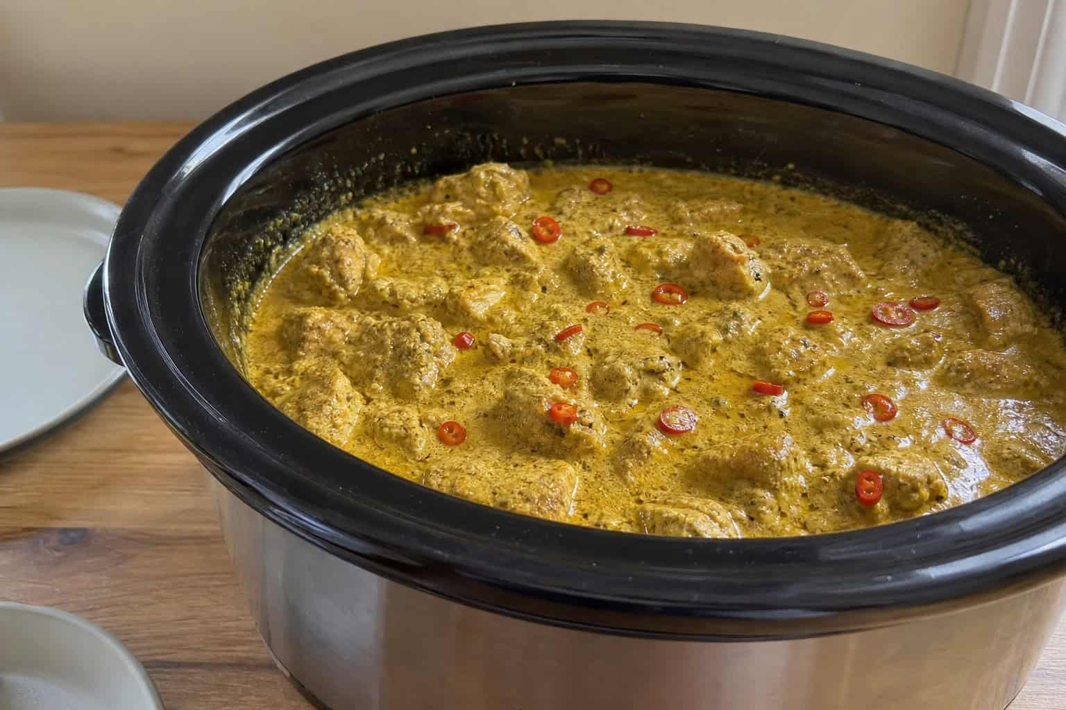 A crockpot filled with coconut curry chicken surrounded by fresh ingredients on a kitchen countertop.