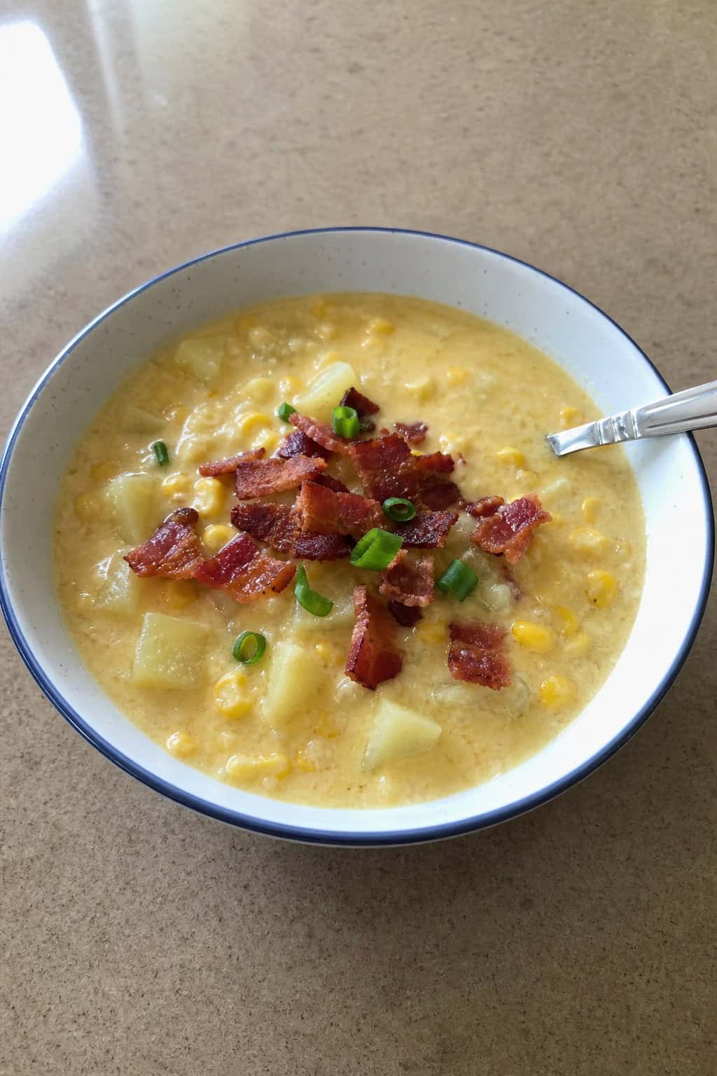 A bowl of creamy corn chowder topped with crispy bacon on a wooden table, surrounded by fresh corn and bacon strips.