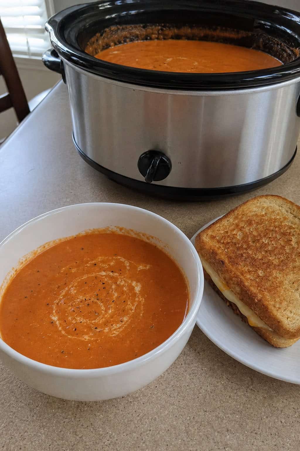 A crockpot filled with creamy tomato basil soup on a wooden countertop surrounded by fresh tomatoes, basil, and garlic.