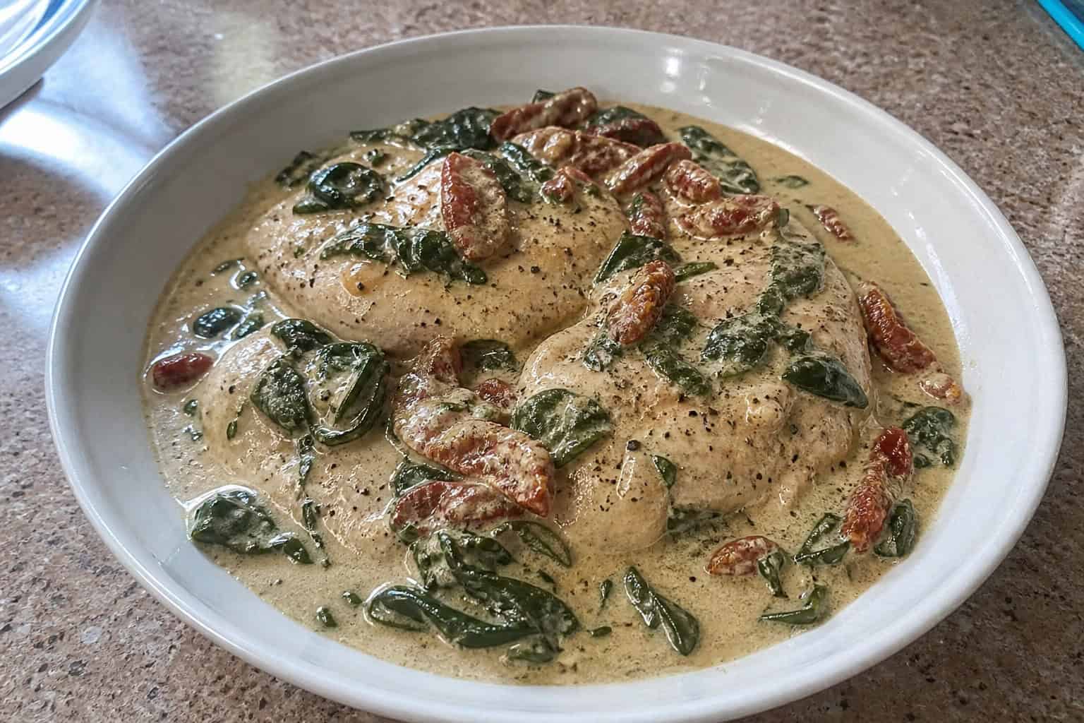 27 Crockpot Chicken Recipes for Effortless Weeknight Dinners 26 A bowl of creamy Tuscan chicken with spinach and sun-dried tomatoes on a wooden table surrounded by fresh ingredients.