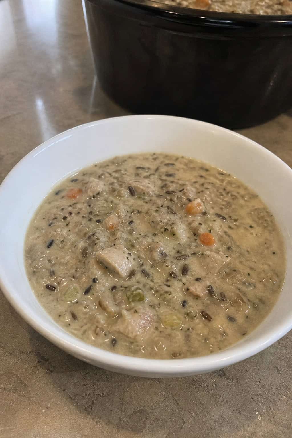 A crockpot filled with creamy wild rice and chicken soup on a wooden countertop surrounded by fresh vegetables and herbs.
