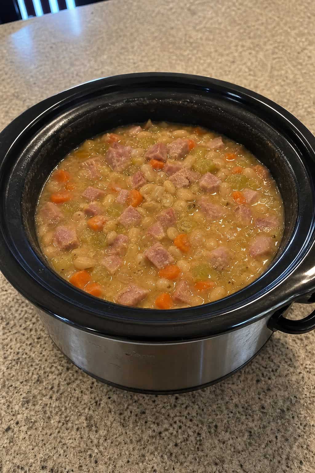 A crockpot filled with ham and bean soup with vegetables on a kitchen countertop surrounded by fresh ingredients.
